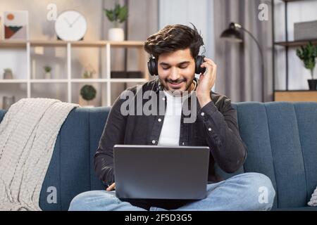 Online lessons, e-learning concept. Young likable Indian man sitting on couch in living room at home, enjoying studying using laptop and headset, looking at screen and listening audio lessons. Stock Photo