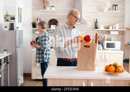 Elderly husband and wife taking out vegetables from grocery paper bag after arriving from supermarket. Cheerful happy family healthy lifestyle putting fresh fruits and groceries in refrigerator Stock Photo