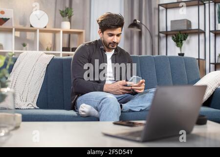 People, home and using gadgets. Young bearded Indian man, sitting on the couch at cozy living room indoors and using smartphone for typing message or serfing social networks. Stock Photo