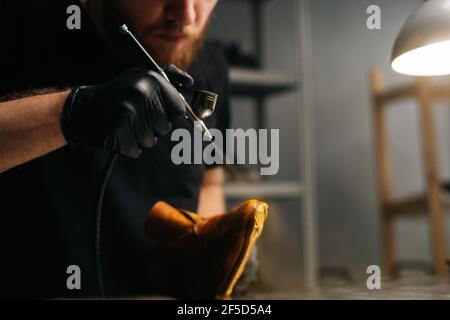 Closeup view of bearded shoemaker wearing black gloves spraying paint of light brown leather shoes, close-up. Stock Photo