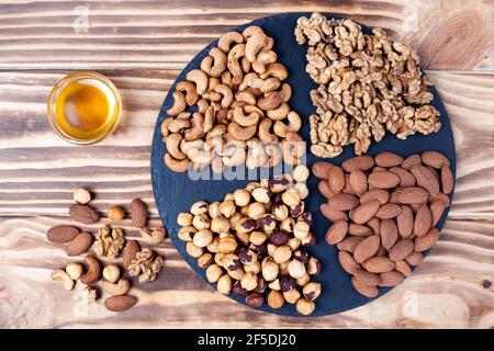 Various nuts sorted on stone plate with honey glass bowl. Mixed nuts on wooden table. Black stone plate on wooden background. Stock Photo