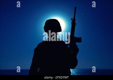 Silhouette of army soldier standing with service rifle in hand on background of night sky with moon. Armed Marines infantry in battle helmet and uniform ready for action, patrolling area in darkness Stock Photo