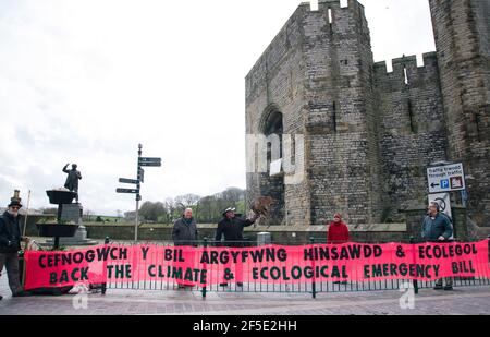 Caernarfon, Gwynedd, North Wales, UK. 26th March 2021. Members of Extinction Rebellion North Wales joined by Plaid Cymru MP Hywel Williams hold a banner calling for the backing of the Climate and Ecological Emergency bill outside Caernarfon Castleas part of a national banner drop day of action.Parliament declared a Climate Emergency back in 2019 – but protestors say actions haven’t matched their words. An emergency requires strong, decisive action to reverse the climate and ecological crisis. Stock Photo