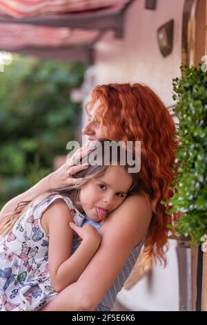 A beautiful red-haired young mother gently holds, hugs little daughter in arms near house with trailer truck. The girl smiles, shows tongue to the cam Stock Photo