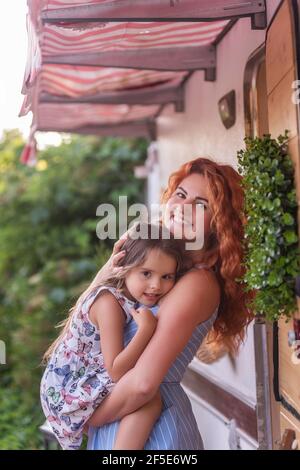 A beautiful red-haired young mother gently holds, hugs little daughter in arms near house with trailer truck. The girls smiles. Cheerful, care, happy Stock Photo