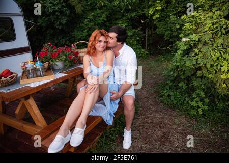 A young brunette man gently kisses a beautiful red-haired girl on the forehead, she laughs. Close-up portrait of couple in love, resting on picnic at Stock Photo