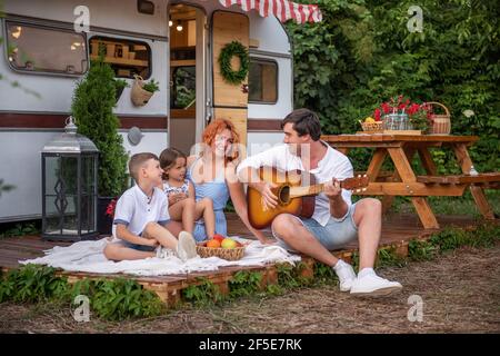 Young father plays the guitar, red-haired mother with children sing songs. Happy Family on picnic, camping by trailer truck in nature outside the city Stock Photo