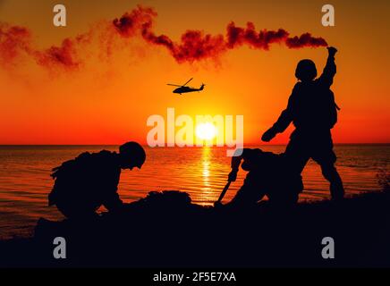 Silhouette of army special forces infantry soldiers, Marines or Navy SEALS team signaling to helicopter with smoke flair while waiting for evacuation, landing on seashore during amphibious operation Stock Photo