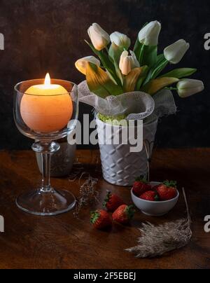 A round candle is lit in a glass candle holder near a vase of white flowers on a wooden table with some strawberries and dry plants Stock Photo