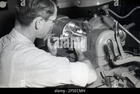 1950s, historical, precision engineering, intricate work as a skilled male worker sitting at bench lathe holds in place metal hand tools in place on the object being cut by the mechanical lathe in the process of making a smal specialist part, possibly a glass lens. A lathe is a rotating machine tool used for various complex engineering tasks involving cutting and grinding metal and other materals. The history of developement of industrial lathes is a fascinating one Stock Photo