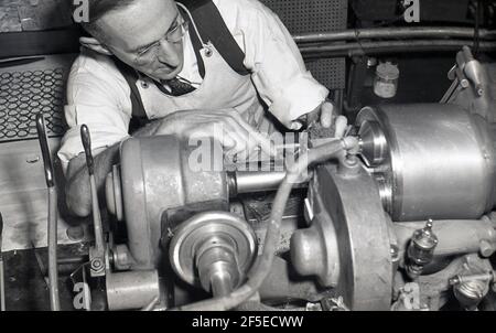 1950s, historical, precision engineering, intricate work as a skilled male worker holds a small wet coth in one hand and with the other holds the object in place, as it is cut by a mechanical tool, an industrial lathe, in the process of making a smal specialist part, in this case possibly a glass lens. A rotating machine tool, a lathe us used to make intricate parts. Seen here is a screw machine or in Britain, a capstan lathe, used for the precision finishing of parts or in an intermediate stage of the produciton process. Stock Photo