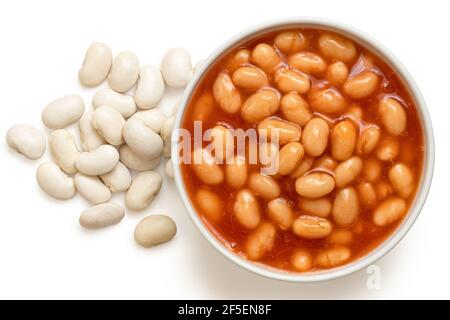 Baked beans in tomato sauce in a white ceramic bowl next to uncooked haricot beans isolated on white. Top view. Stock Photo