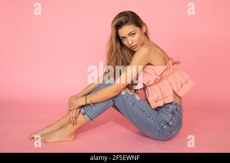 Pretty young woman in pink shirt and jeans sitting on floor on pink background in studio barefoot, Stock Photo