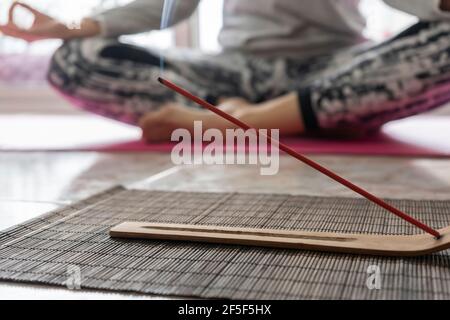 Woman practicing yoga.Focus on burning incense stick with smoke.Yoga lifestyle at home, concept of girl meditating in background. Stock Photo
