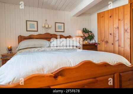 Queen size bed with solid pine wood-frame, headboard, footboard in upstairs master bedroom with white painted narrow wood plank walls inside old house Stock Photo