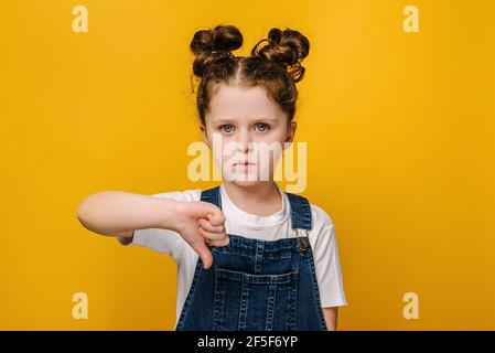 Portrait of serious little girl look at camera show thumb down gesture symbol of negative feedback and dislike, isolated over yellow studio wall Stock Photo