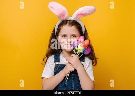 Good looking smiling preschool girl kid with bunny fluffy ears, holds painted eggs on sticks, has a happy look, going to celebrate Easter Stock Photo