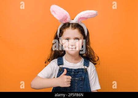 Portrait of happy charming little girl kid showing thumb up as sign of approval or agreement isolated over orange studio background Stock Photo