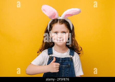 Close up head shot portrait of smiling little girl in pink bunny fluffy ears. Happy kid liking and thumb up on yellow studio background Stock Photo