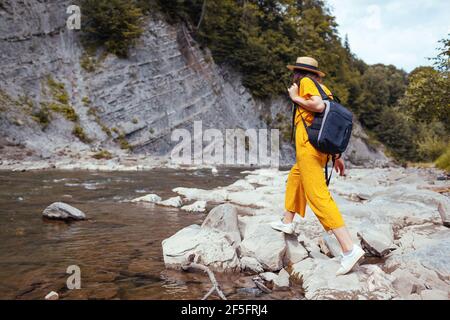 Happy tourist woman hiking by mountain river enjoying landscape. Traveler walking with backpack along rocks. Summer trip close to nature Stock Photo