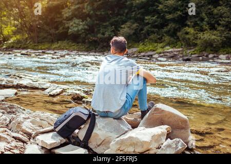 Man tourist relaxing by mountain river enjoying natural landscape. Traveler backpacker sitting on rock. Summer tour to Carpathians Stock Photo