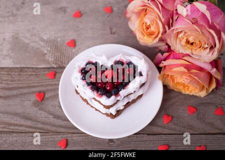 Mini romantic dessert cake for Valentine's Day with roses. Sweet cookies with cream topping and red heart for decor on wooden background. Close-up, co Stock Photo