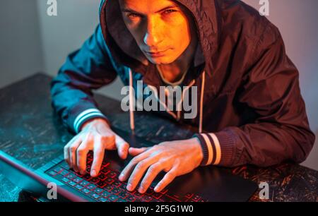 A hooded hacker is typing on a laptop keyboard in a dark room under a neon light. The concept of cybercrime fraud and identity theft Stock Photo