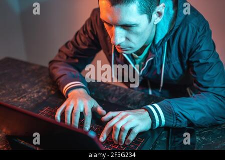 A male hacker is typing on a laptop keyboard in a dark room under a neon light. The concept of cybercrime fraud and identity theft Stock Photo