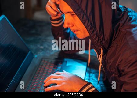 A hooded hacker is typing on a laptop keyboard in a dark room under a neon light. The concept of cybercrime fraud and identity theft Stock Photo