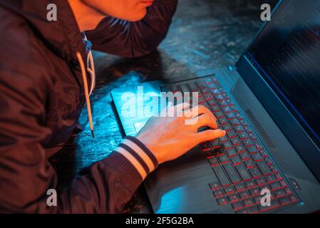 A hooded hacker is typing on a laptop keyboard in a dark room under a neon light. The concept of cybercrime fraud and identity theft Stock Photo