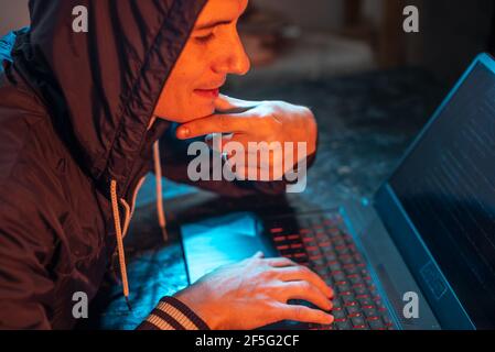 A hooded hacker is typing on a laptop keyboard in a dark room under a neon light. The concept of cybercrime fraud and identity theft Stock Photo