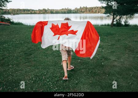 Girl wrapped in large Canadian flag by Muskoka lake in nature. Canada Day celebration outdoors. Kid in large Canadian flag celebrating national Canada Stock Photo