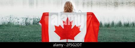 Girl wrapped in large Canadian flag by Muskoka lake in nature. Canada Day celebration outdoors. Kid in large Canadian flag celebrating national Canada Stock Photo