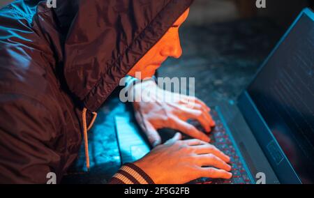 A hooded hacker is typing on a laptop keyboard in a dark room under a neon light. The concept of cybercrime fraud and identity theft Stock Photo