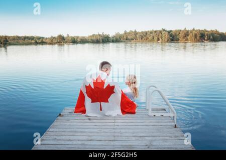 Father and daughter wrapped in large Canadian flag sitting on wooden pier by lake. Canada Day celebration outdoors. Dad and child sitting together Stock Photo