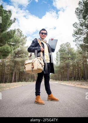 Shallow focus of a young stylish man with a bag and a laptop standing on the road in a forest Stock Photo