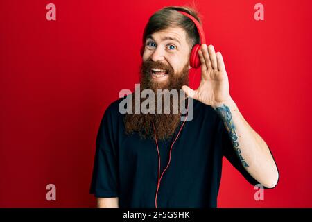 Redhead man with long beard listening to music using headphones smiling with hand over ear listening an hearing to rumor or gossip. deafness concept. Stock Photo