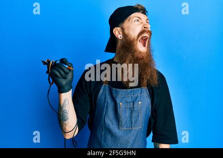 Redhead man with long beard tattoo artist wearing professional uniform and gloves angry and mad screaming frustrated and furious, shouting with anger. Stock Photo