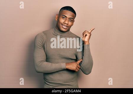 Young black man wearing casual turtleneck sweater with a big smile on face, pointing with hand and finger to the side looking at the camera. Stock Photo