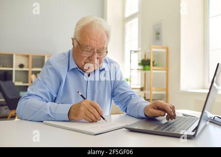 Senior man in eyeglasses sitting at desk at home, using laptop and taking notes in notebook Stock Photo