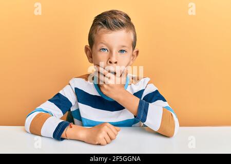 Adorable caucasian kid wearing casual clothes sitting on the table looking fascinated with disbelief, surprise and amazed expression with hands on chi Stock Photo