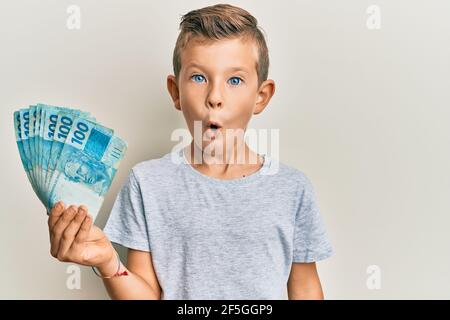 Adorable caucasian kid holding 100 brazilian real banknotes scared and amazed with open mouth for surprise, disbelief face Stock Photo