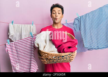 Handsome hispanic man doing laundry holding wicker basket making fish face with mouth and squinting eyes, crazy and comical. Stock Photo