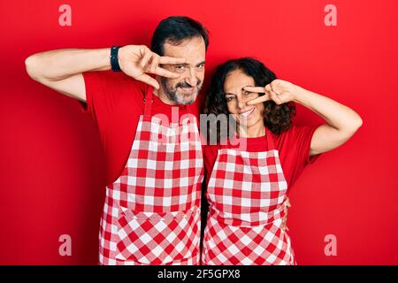 Middle age couple of hispanic woman and man wearing cook apron doing peace symbol with fingers over face, smiling cheerful showing victory Stock Photo