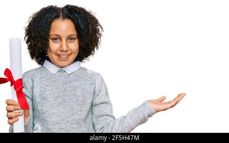 Young little girl with afro hair holding graduate degree diploma celebrating victory with happy smile and winner expression with raised hands Stock Photo