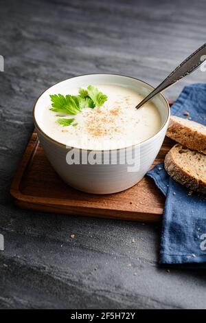 Delicious creamy celery soup topped with black pepper and fresh leaves, served with crusty bread, on rustic stone background Stock Photo