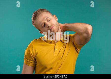Tired young man having pain - neck ache. Guy in casual yellow t-shirt takes break and kneads sore neck muscles with hand massage. Stock Photo