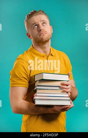 European student is dissatisfied with amount of homework and books on blue background. Man sighs and rolls his eyes in displeasure, he is annoyed Stock Photo