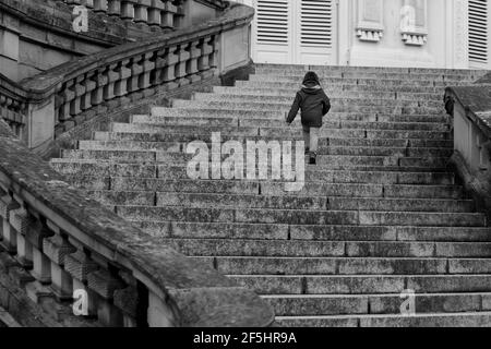 Unrecognisable little girl, modern-day lonely princess walking up majestic curved stairs of empty castle in Europe. Black and white low angle Stock Photo