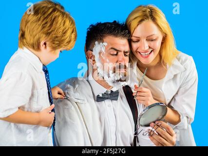 Hairdresser making hairstyle. Family day. Mother cutting hair to father and little son shaving dad beard with razor. Stock Photo